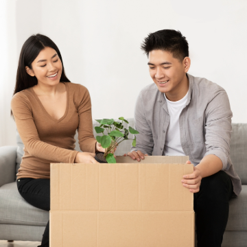 how-to-save-for-a-house_landing-page.png Happy man and happy woman unpacking green plant from cardboard box in new home