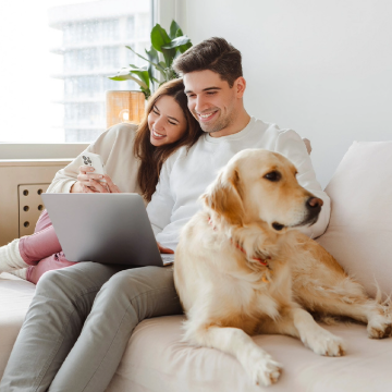 prioritizing-financial-health_tile-size.png Happy couple sitting couch working on laptop next to white dog